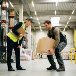 Nederlandse arbeidsinspecteur observeert werknemer die correct een zware doos tilt in moderne magazijn met veiligheidsapparatuur