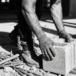 Bouwvakker grijpt betonblokken vast, gespannen spieren en zweet zichtbaar op verweerde onderarmen op bouwplaats