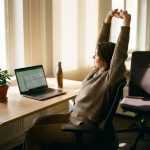 Persoon strekt armen boven hoofd tijdens werk aan bureau met laptop, ergonomische stoel en plant in natuurlijk licht