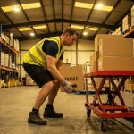 Magazijnmedewerker in veiligheidsvest tilt pakket met juiste hefttechniek naast hydraulische heftafel in industrieel pakhuis