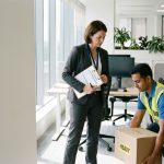 Veiligheidsinspecteur met klembord observeert werknemer die juiste tiltechniek demonstreert in modern kantoor