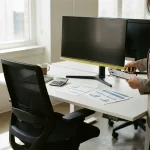 Ergonomiespecialist meet monitorhoogte aan bij modern bureaustation met meetlint en klembord op wit bureau