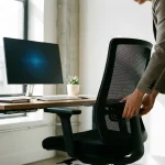 Kantoormedewerker past ergonomische bureaustoel aan, handen op lumbaalsteun, moderne werkplek met monitor en plant