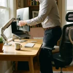 Kantoormedewerker past ergonomische monitorarm aan zittend aan georganiseerd bureau met laptop en plant bij raam