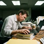 Kantoormedewerker met slechte houding achter laptop, gebogen nek en opgetrokken schouders, rommelig bureau