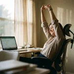Kantoormedewerker strekt armen boven hoofd aan ergonomisch bureau met laptop, plant en natuurlijk licht door raam