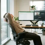 Persoon doet nekrek met handen achter hoofd aan ergonomisch bureau met verstelbare stoel en meerdere monitoren