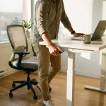 Kantoormedewerker staat op bij verstelbaar zit-sta bureau met laptop en koffie in moderne werkruimte met natuurlijk licht